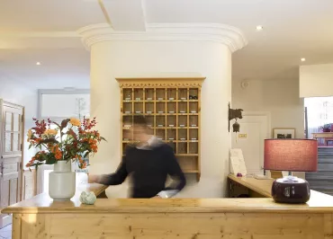 Réception d'hôtel lumineuse et accueillante. Derrière un grand comptoir en bois clair, on peut apercevoir une personne souriante. Des fleurs colorées dans un vase apportent une touche de fraîcheur à l'espace.