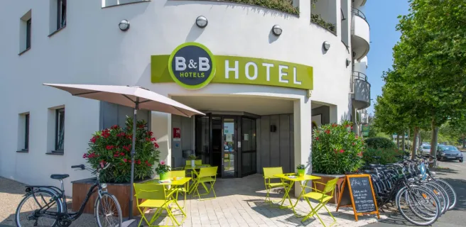 La facade extérieure de l"hôtel, avec des tables et chaises métalliques vertes devant.